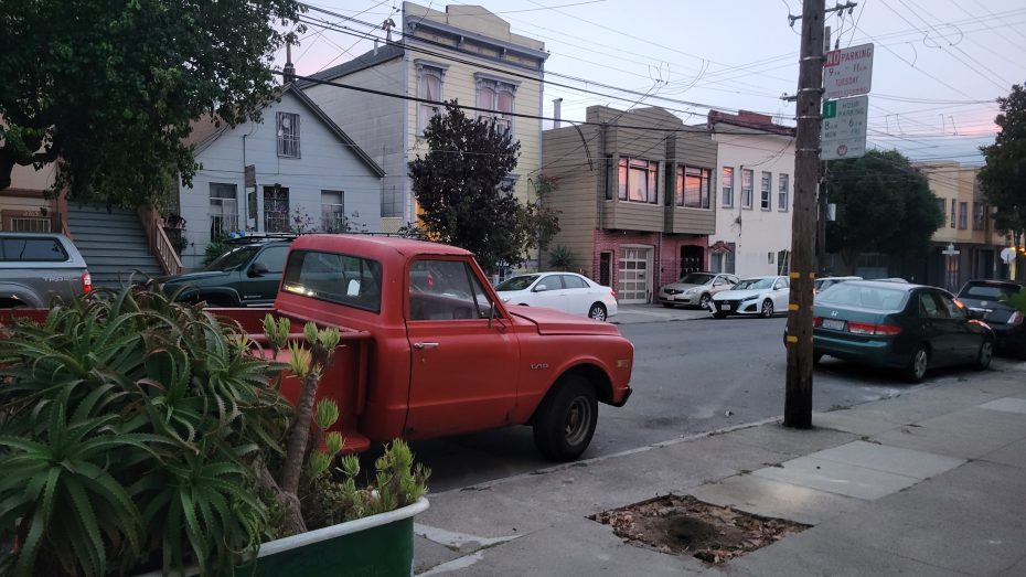 A red vintage pickup truck is parked on a residential street lined with houses. Other cars are parked along the street, and power lines are visible overhead.