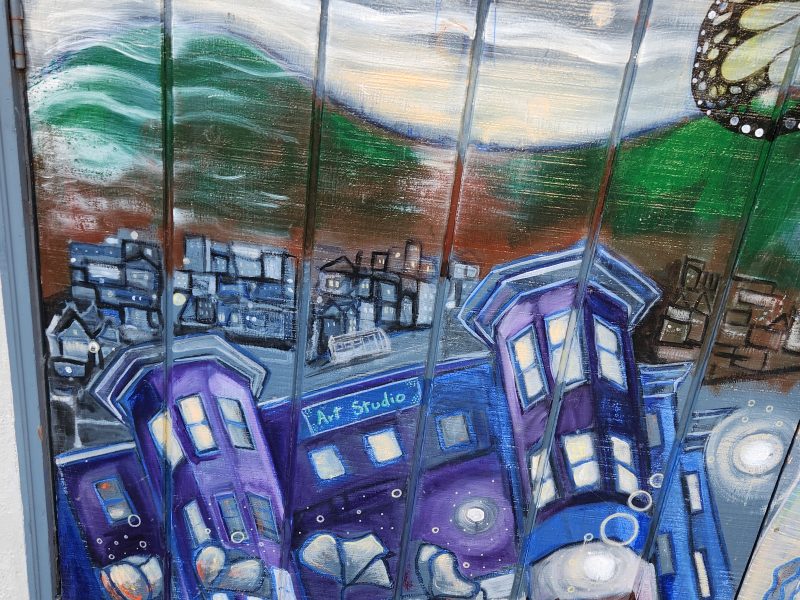 Colorful mural depicting a cityscape with purple buildings, mountains, and a butterfly in the corner. The words "Art Studio" are visible on one building.