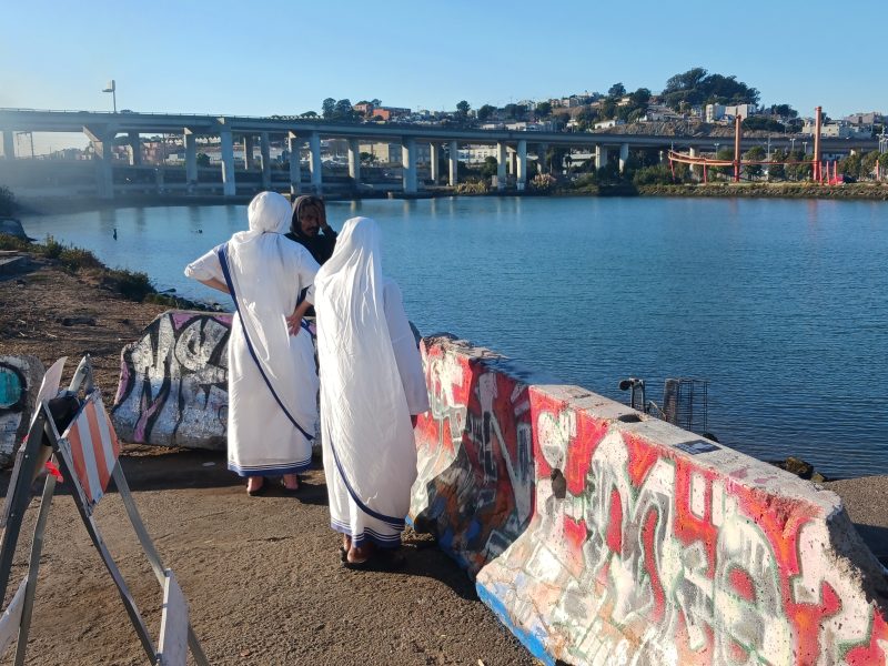 Three people in white garments stand near a graffiti-covered barrier by a body of water, with a highway bridge and hills in the background.