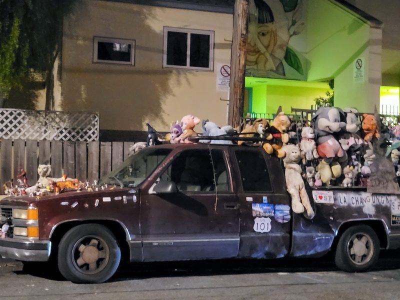 A truck covered in numerous stuffed animals and decals is parked on the street at night.