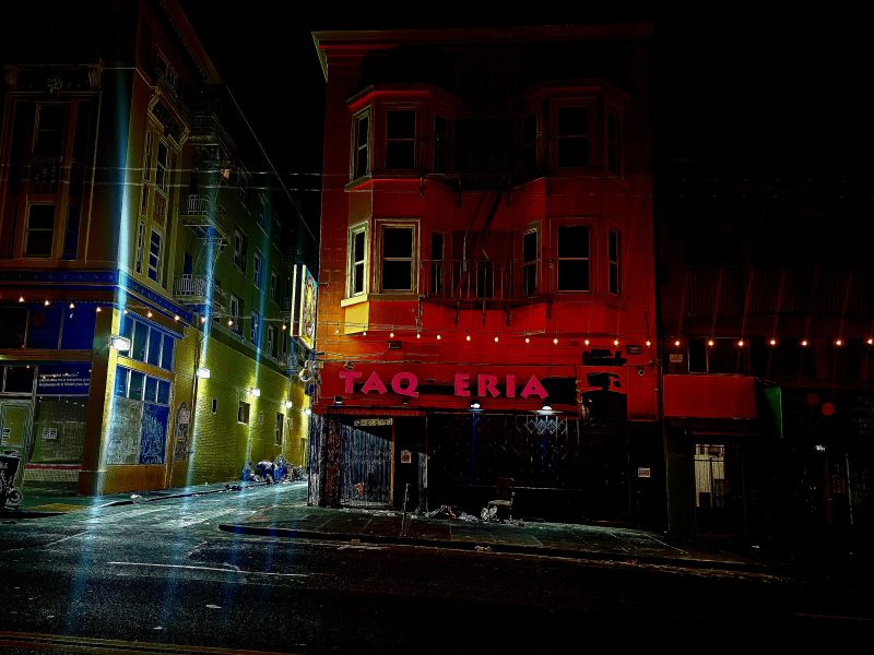 Street view of a taqueria at night, with dim lighting and colorful buildings on each side.