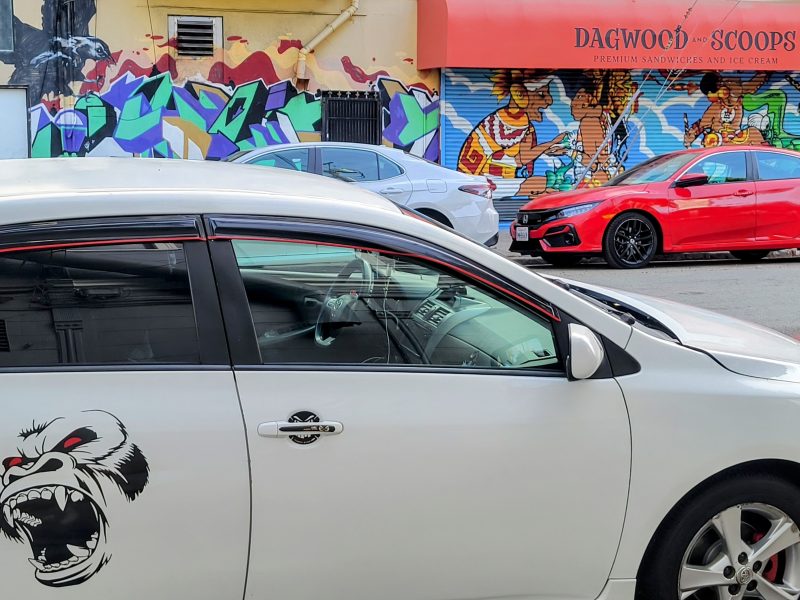 Cars parked on a street with a white car featuring a gorilla decal. A colorful mural is visible behind them. A storefront with an orange awning reads "Dagwood Scoops.