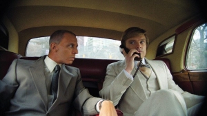 Two men in suits sit in the back of a vintage car. One man is talking on a corded phone, and the other listens attentively. Trees and a building are visible through the windows.