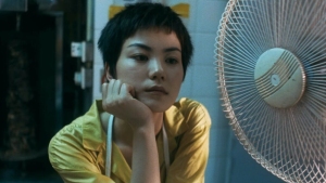 A person with short hair wearing a yellow shirt leans on their hand while looking at a table fan.