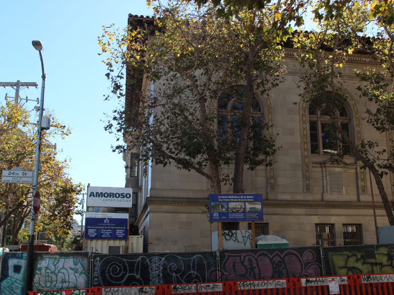 A historic mission library under renovation is surrounded by fences and construction signs, with trees lining the street. Graffiti adorns the barriers, adding a touch of urban art to the scene.