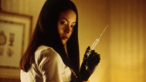 A woman with long dark hair, wearing black gloves, holds a syringe while looking at it in a dimly lit room.