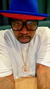 Man wearing a blue and red hat, black glasses, white shirt, and a gold necklace with a pendant, looking at the camera.
