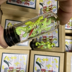 A hand holding a small container with a green toy bug inside. Background shows stacked boxes with labels.