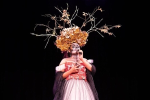 Person in elaborate costume with a floral headpiece and face paint, standing on stage holding a microphone.