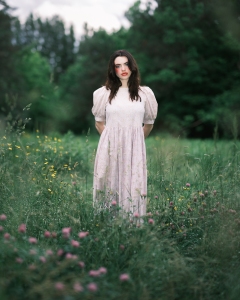 Woman in a white dress stands in a field of tall grass and wildflowers, with a forest in the background.