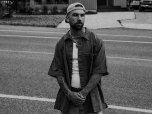 A person stands in the middle of a road, wearing a cap, open shirt, and white t-shirt. The background features pavement and building structures. The image is in black and white.