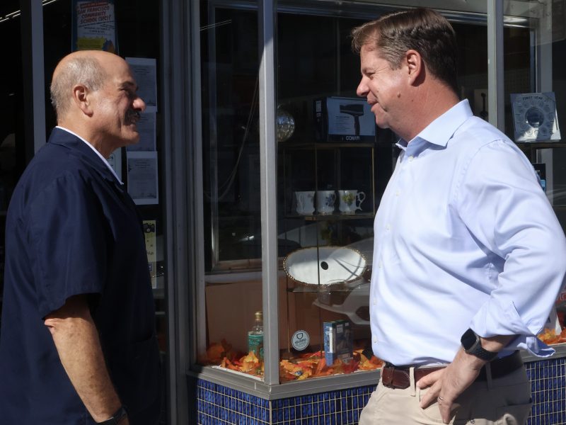 Two men having a conversation outside a store with glass windows. One man is wearing a dark jacket, while the other is in a light blue shirt.