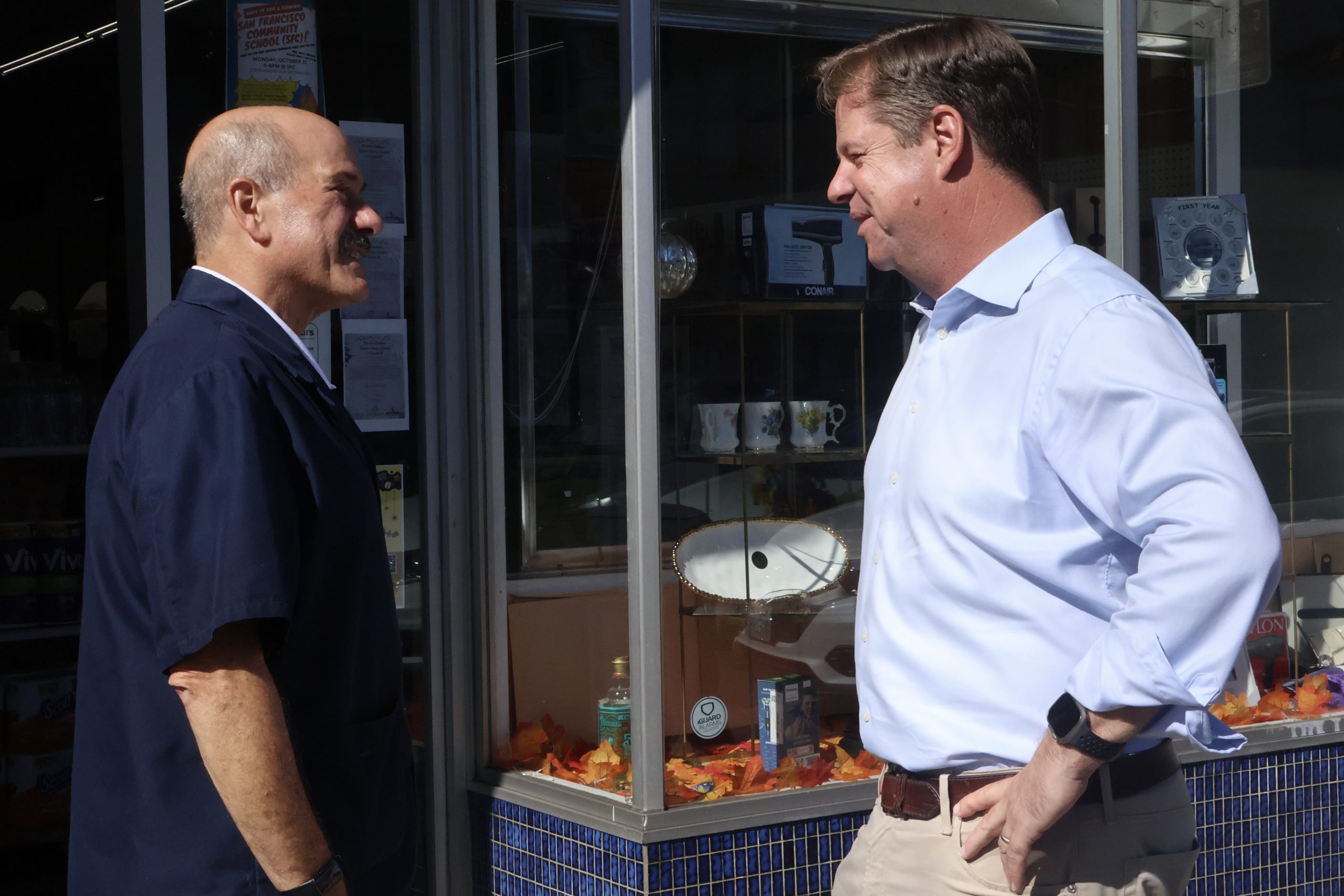 Two men having a conversation outside a store with glass windows. One man is wearing a dark jacket, while the other is in a light blue shirt.