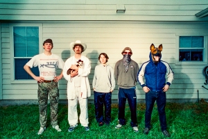 A group of five people stand in front of a house; one holds a dog and another wears a dog mask.