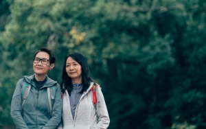 Two people wearing jackets and backpacks stand outdoors against a backdrop of dense green foliage.