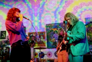 A band performs on stage with colorful, psychedelic art in the background. The vocalist holds a microphone while the guitarist plays. A drummer is visible in the back.