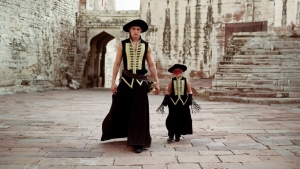 Two people dressed in matching black and gold medieval-style outfits walk through a stone courtyard.