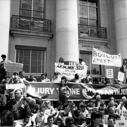A black-and-white photo of a protest outside a building. People hold signs with messages like "Boycott Apartheid," and other banners. A crowd is gathered on the steps and in front of the building.