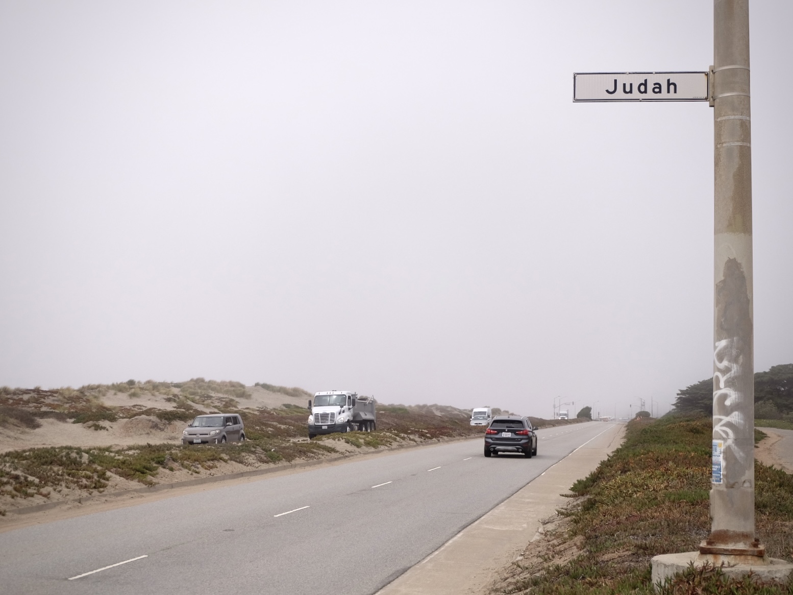 Foggy road with a few vehicles, bordered by dunes and low vegetation. A street sign labeled "Judah" is mounted on a pole.