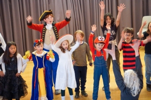 A group of children in costumes with a teacher in front, raising their hands on stage.