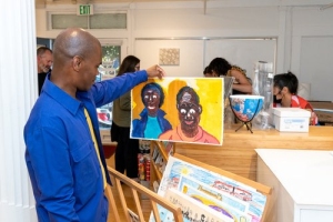A person in a blue shirt holds up a colorful painting of a smiling couple in a room with several people and shelves in the background.