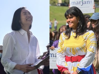 Two women standing outdoors, both smiling. The woman on the left holds a clipboard and wears a white blouse. The woman on the right wears a colorful dress with a sunburst design.