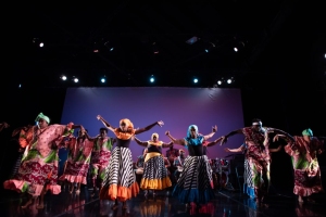 Performers in colorful costumes dance energetically on stage under bright lights.