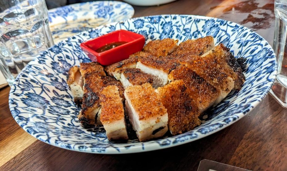 Sliced crispy pork belly on a blue and white patterned plate, served with a small red dish of dipping sauce, on a wooden table.