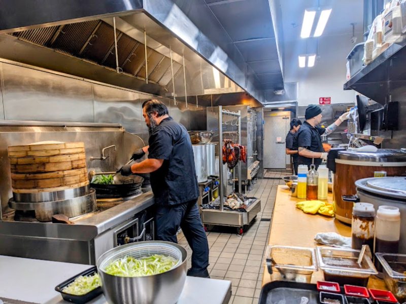 A restaurant kitchen with two chefs cooking. One is stirring a large pot, while the other is at a counter. Various ingredients and pots are visible around the kitchen.