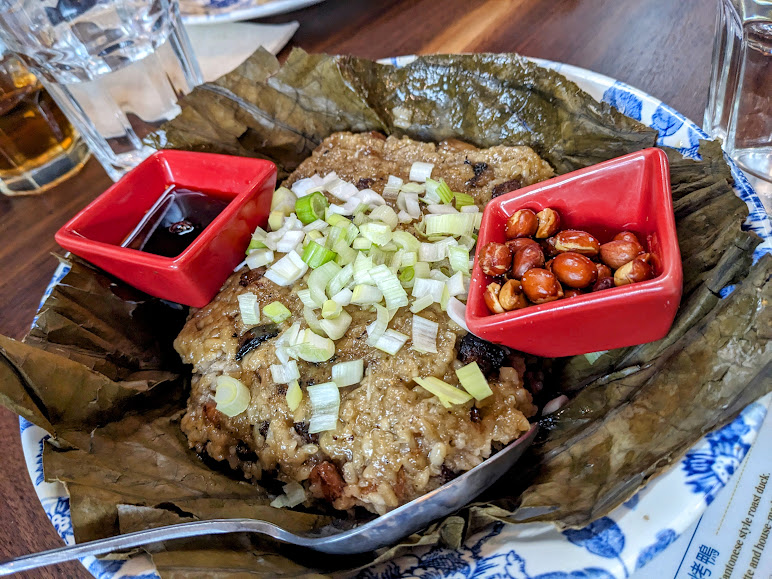 Sticky rice wrapped in a leaf, topped with chopped green onions, served with sauce and peanuts in small red dishes on a patterned plate.