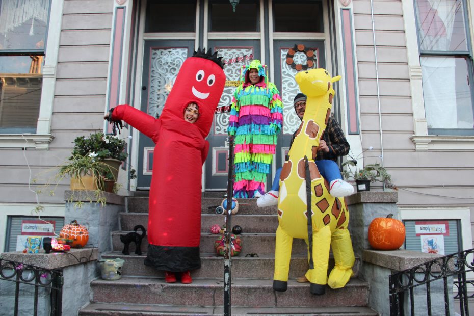 Three people in colorful costumes— a red tube, a rainbow fringe outfit, and a giraffe—stand on a porch with Halloween decorations.