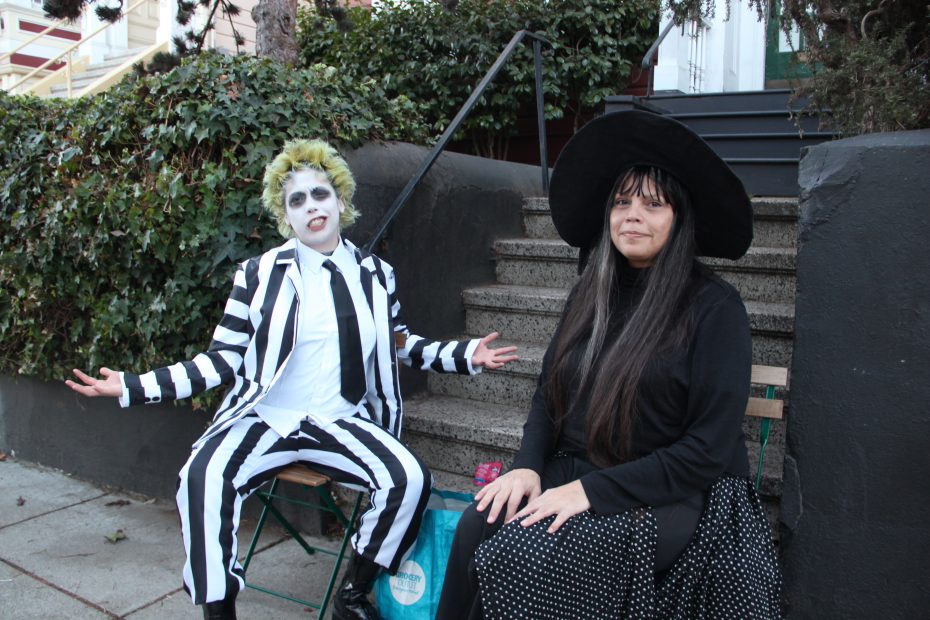 Two people in costume sit outdoors. The left person wears a black and white striped suit and face paint. The right person wears a black dress and large hat.