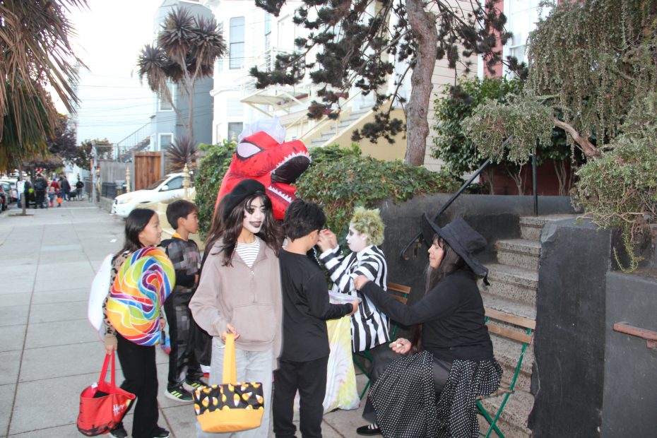 Children in costumes trick-or-treating, with some dressed as a dinosaur, witch, and skeleton, while adults distribute candy on a residential street.