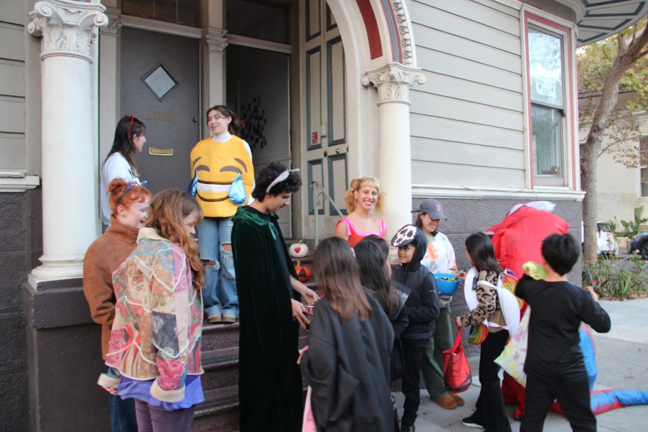 A group of children and adults in costumes gather outside a house with a decorated door. Some hold bags, possibly for Halloween trick-or-treating.