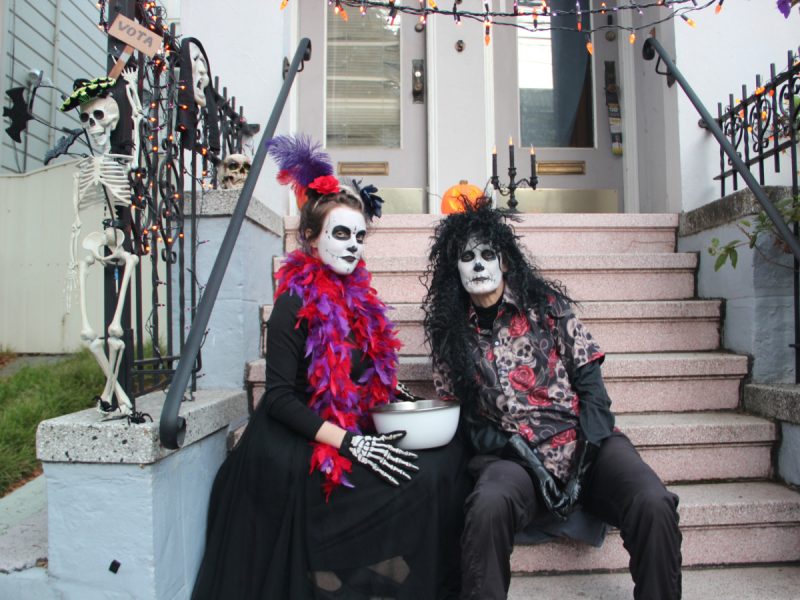 Two people in skeleton costumes sit on stairs decorated with Halloween lights and a skeleton figure. They hold a bowl and are dressed in colorful accessories.
