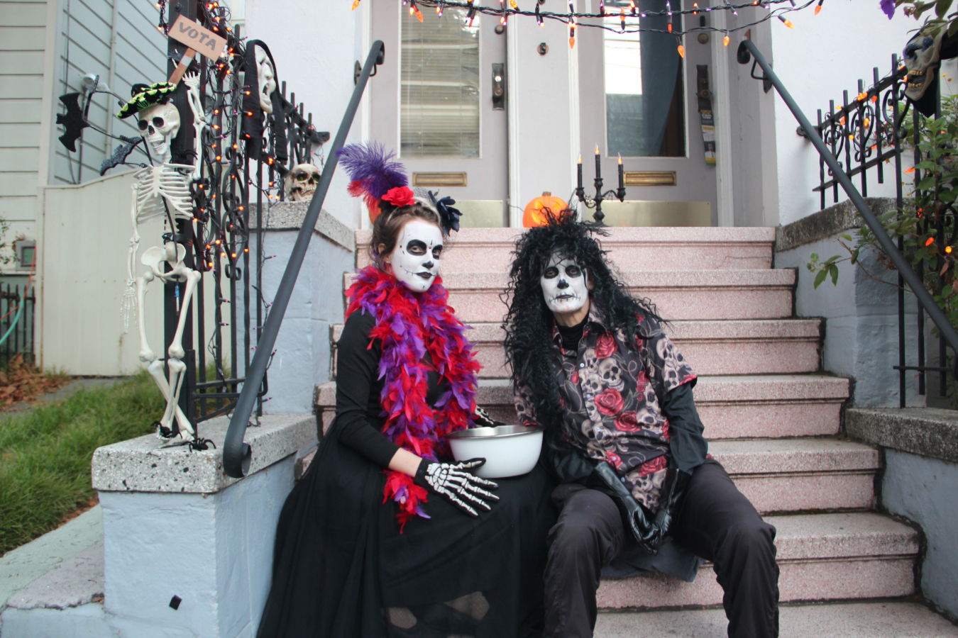 Two people in skeleton costumes sit on stairs decorated with Halloween lights and a skeleton figure. They hold a bowl and are dressed in colorful accessories.