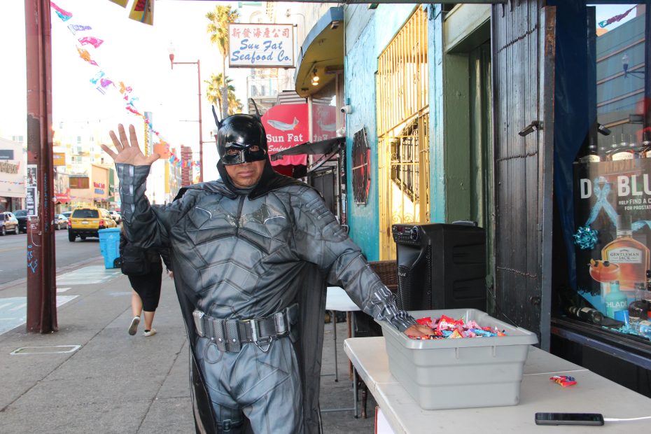 A person dressed as Batman waves near a table with a container of candy on a city street.