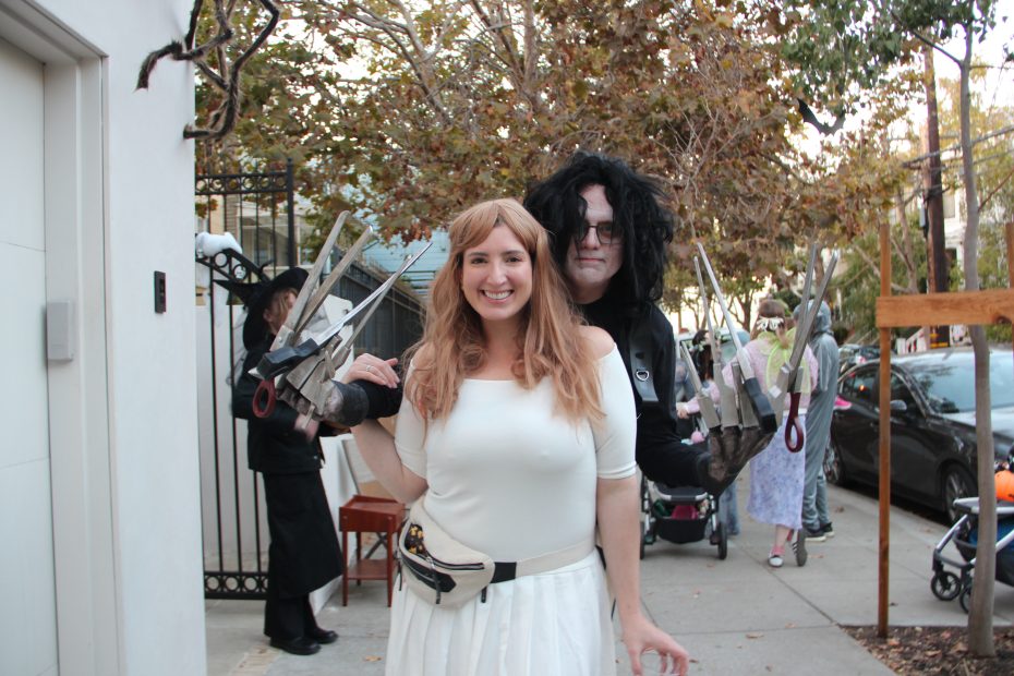A woman dressed as an angel poses in front of a person wearing a scissorhands costume on a tree-lined street.