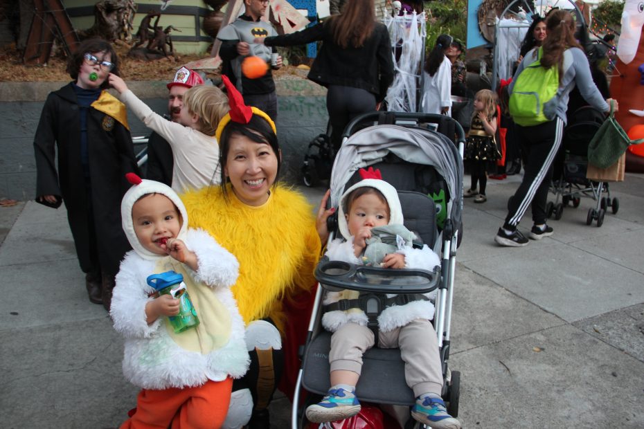 A person and two children in chicken costumes pose for a photo at a Halloween event, surrounded by other people in various costumes.