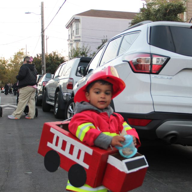 Young child dressed as a firefighter with a red fire truck costume, walking on a street lined with parked cars.