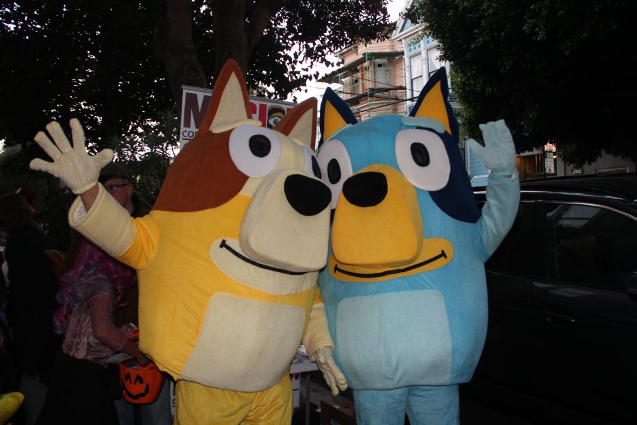 Two people wearing large dog costumes, one brown and the other blue, are standing outdoors with trees and buildings in the background.