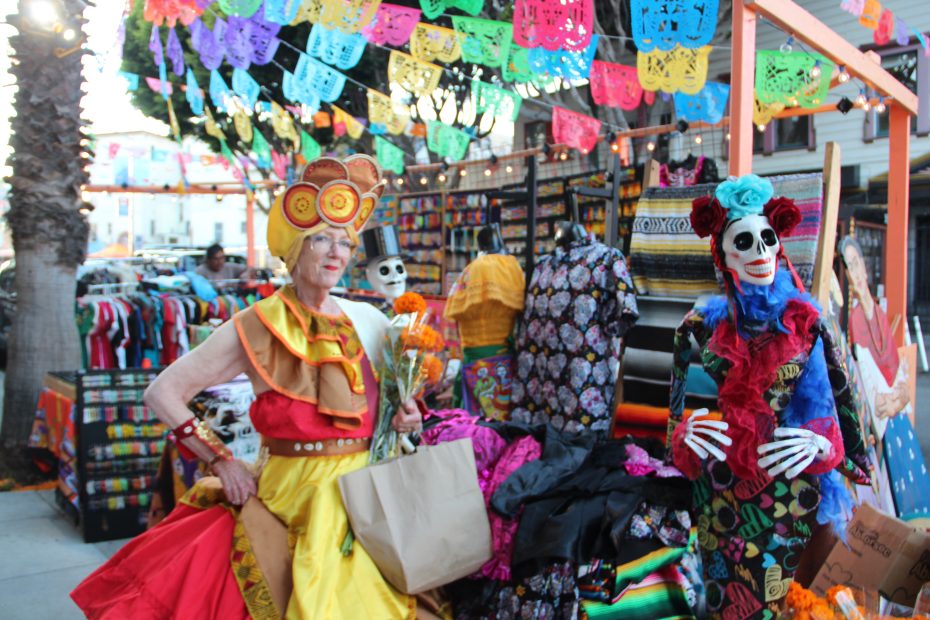 A woman in colorful traditional attire stands beside a skeleton figure in a vibrant market stall, decorated with papel picado and fabrics.