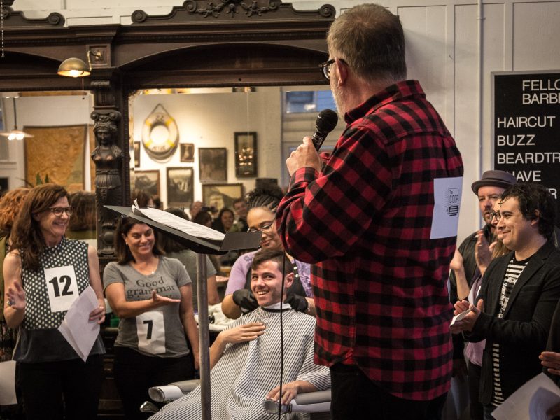 A man in a barber chair holds a microphone, surrounded by a group of people with number cards. A sign in the background lists haircut prices.