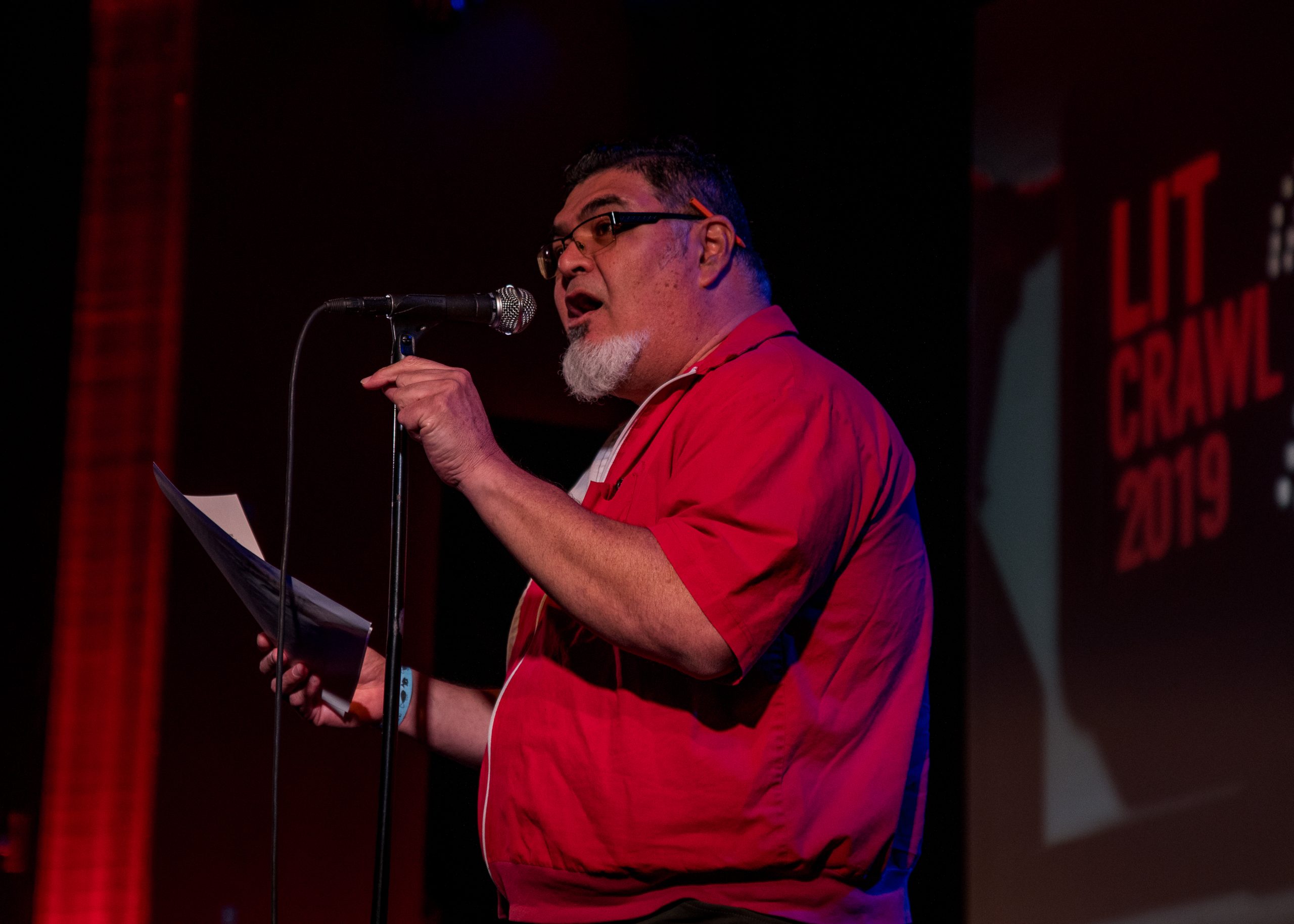 Person in a red shirt speaking into a microphone on stage at an event with "Lit Crawl 2019" on a nearby screen.