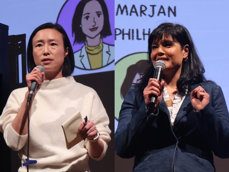 Two women speaking into microphones on stage. One holds notes and is wearing a white sweater; the other is in a dark jacket. A projection with illustrations and text is seen in the background.