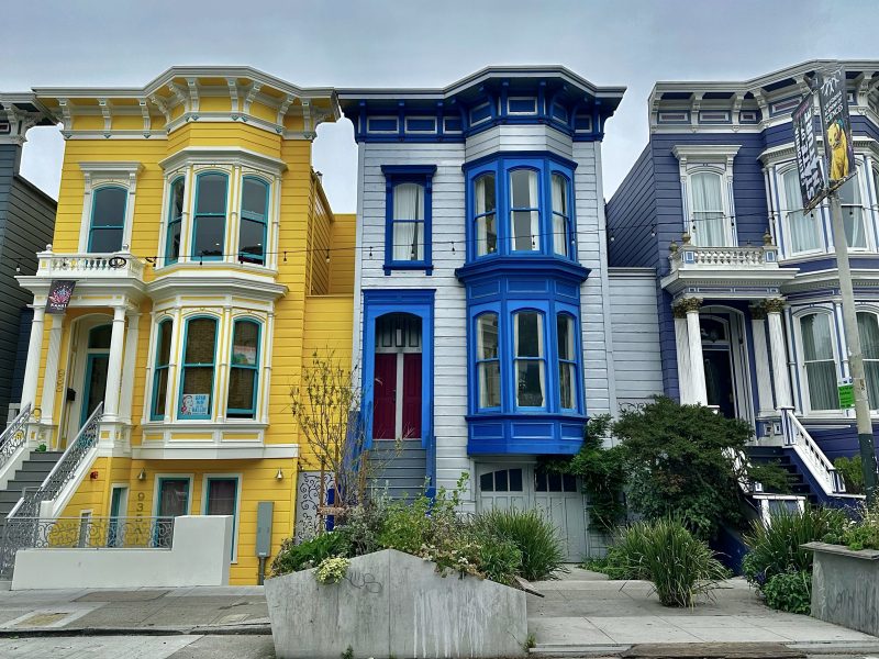 Three colorful Victorian houses in a row, painted yellow, blue, and purple, with detailed architectural features and small front gardens.
