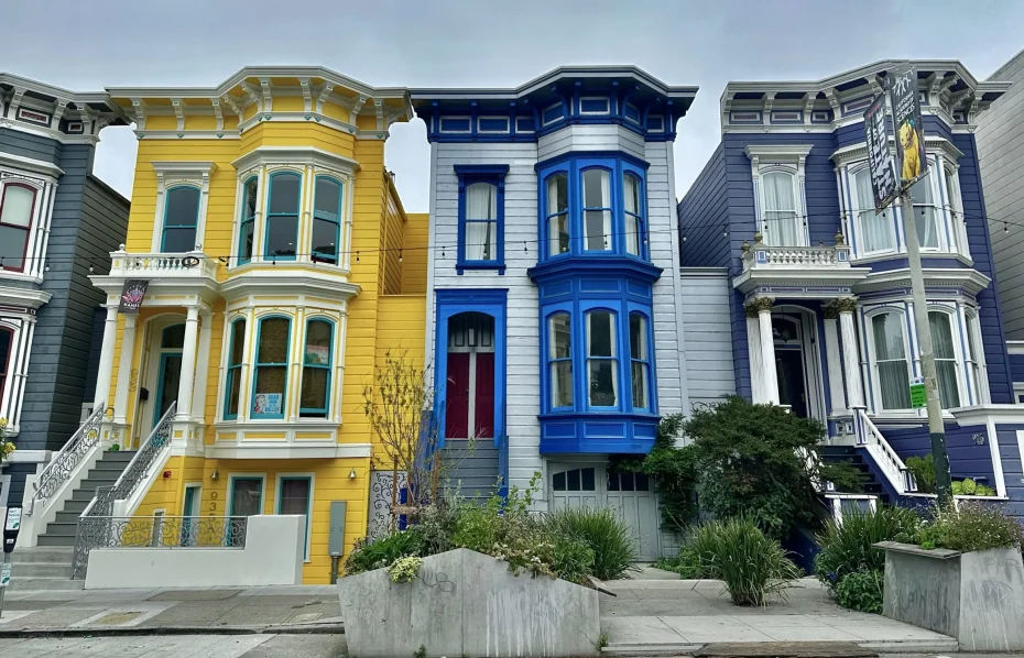 Three colorful Victorian-style houses are lined up: one yellow, one blue, and one dark blue, with a garden in front.