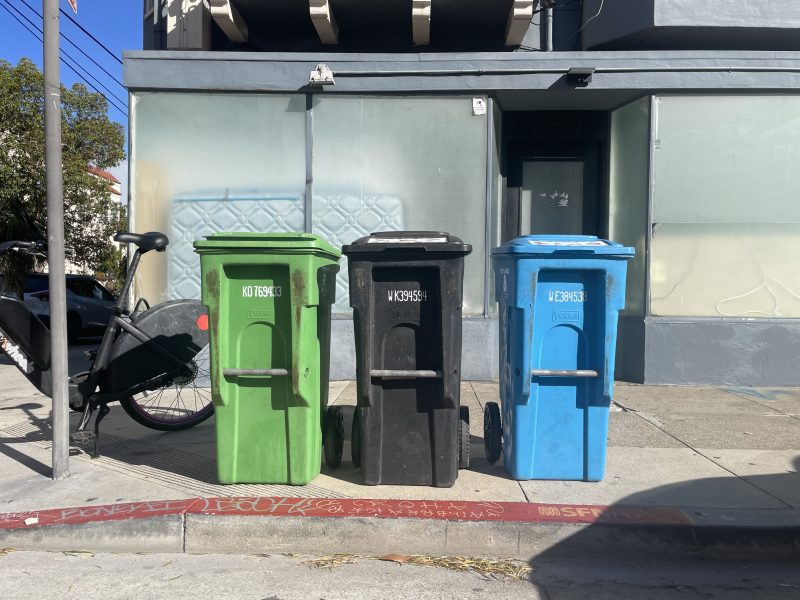 Three wheeled bins—green, black, and blue—are lined up on a sidewalk