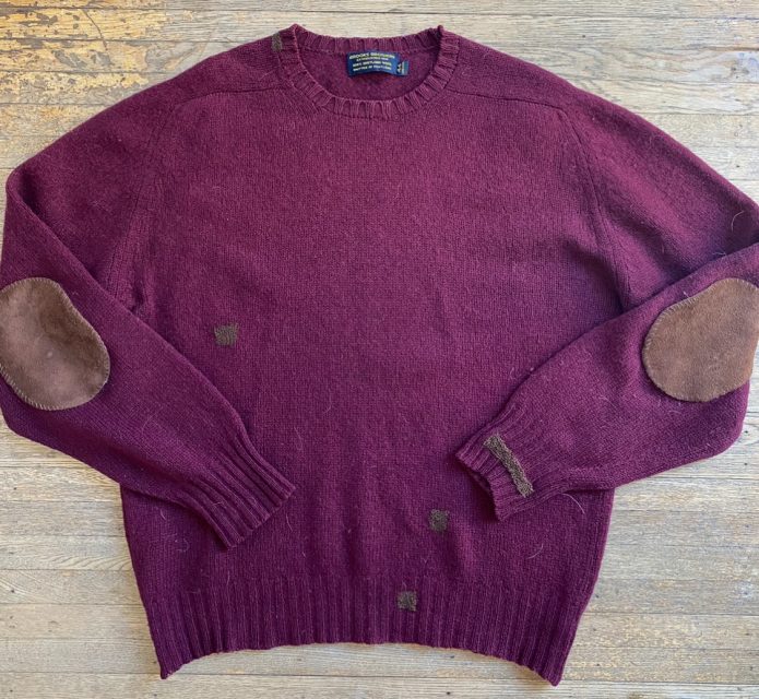 A burgundy knit sweater with brown patches on the elbows and small dark areas throughout, laid flat on a wooden surface.