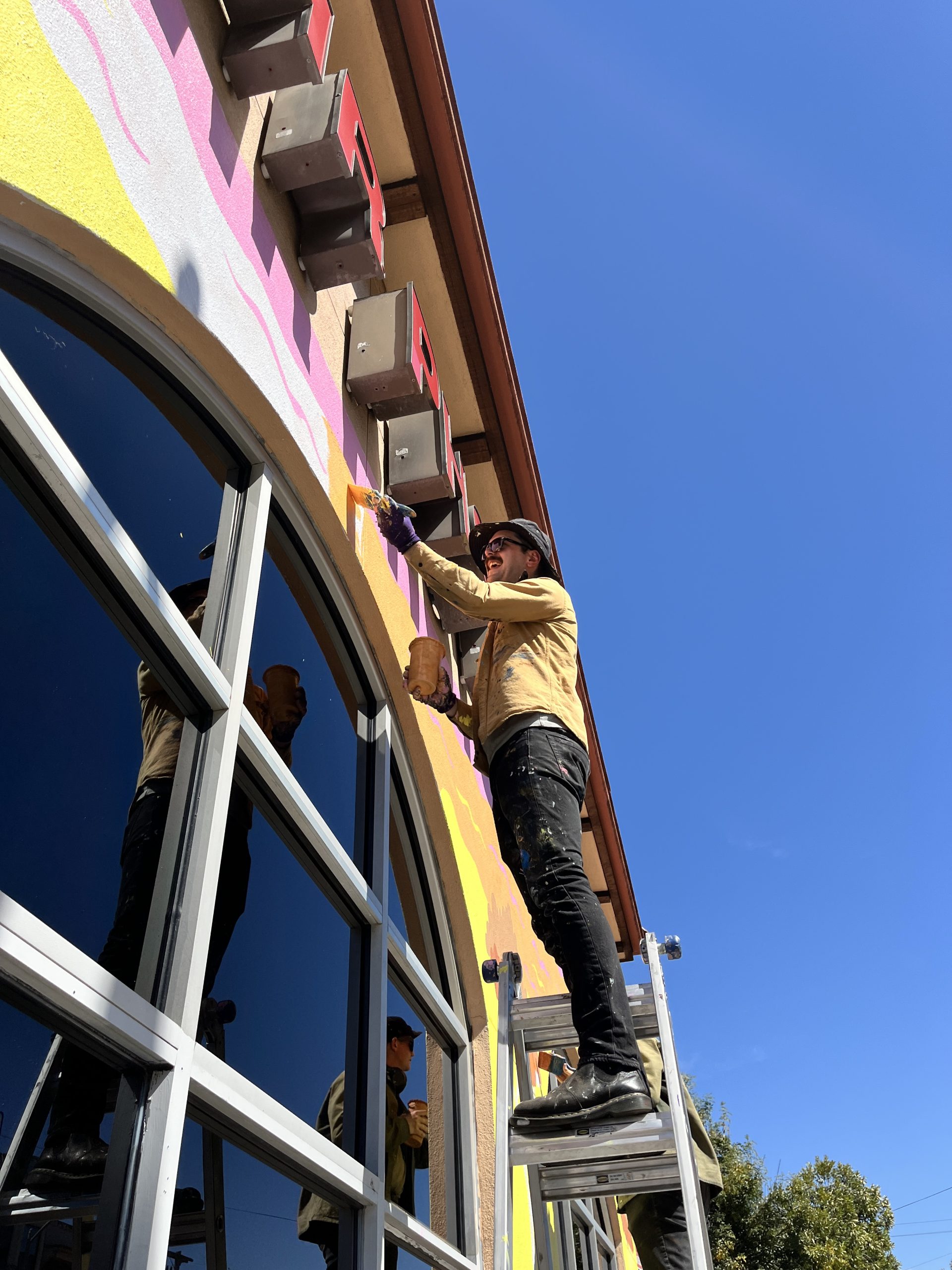 A person on a ladder paints a building's exterior under a clear blue sky.